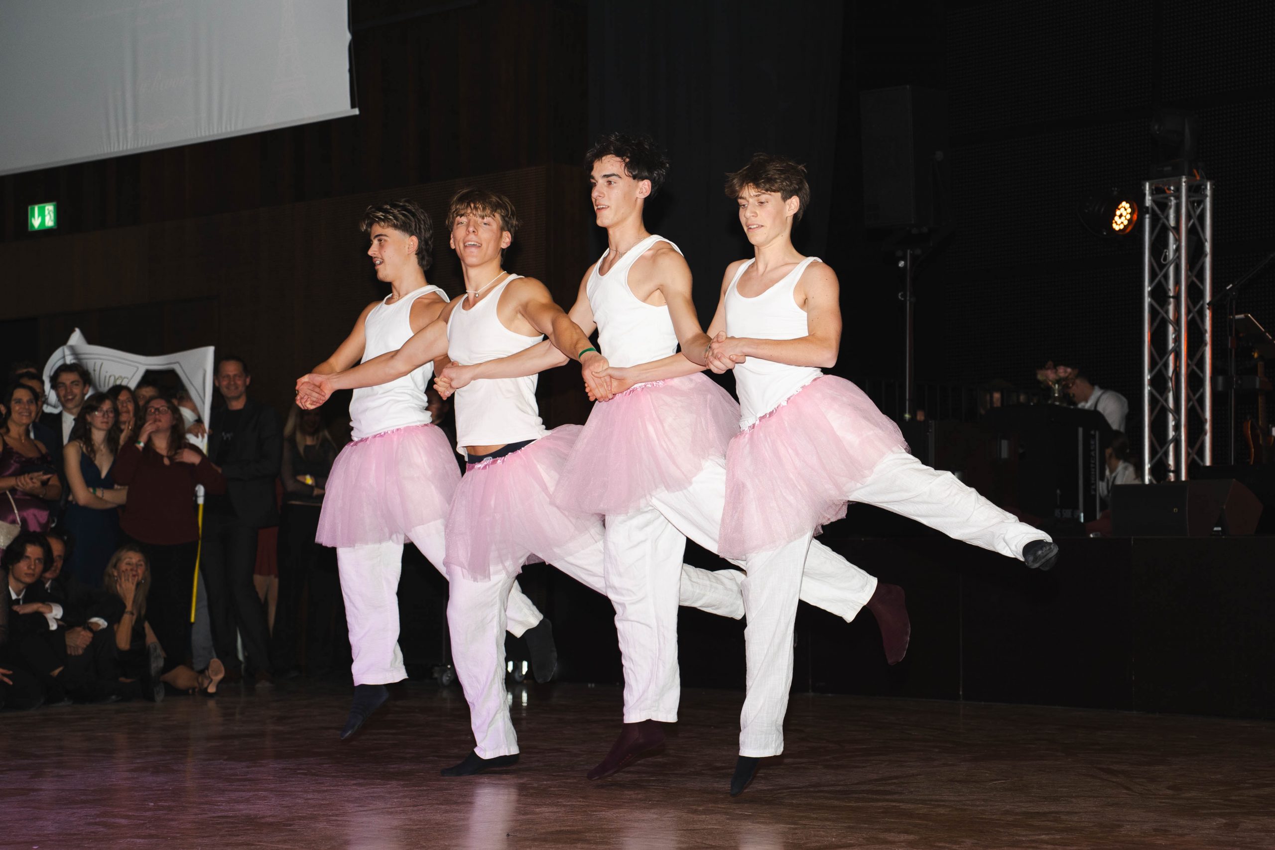 Vier junge Männer in weißen Tanktops, weißen Hosen und rosa Tutus führen auf der Bühne einen synchronen Balletttanz auf, wobei sie ein Bein heben und sich an den Händen halten. Das Publikum schaut zu und lächelt im Hintergrund.