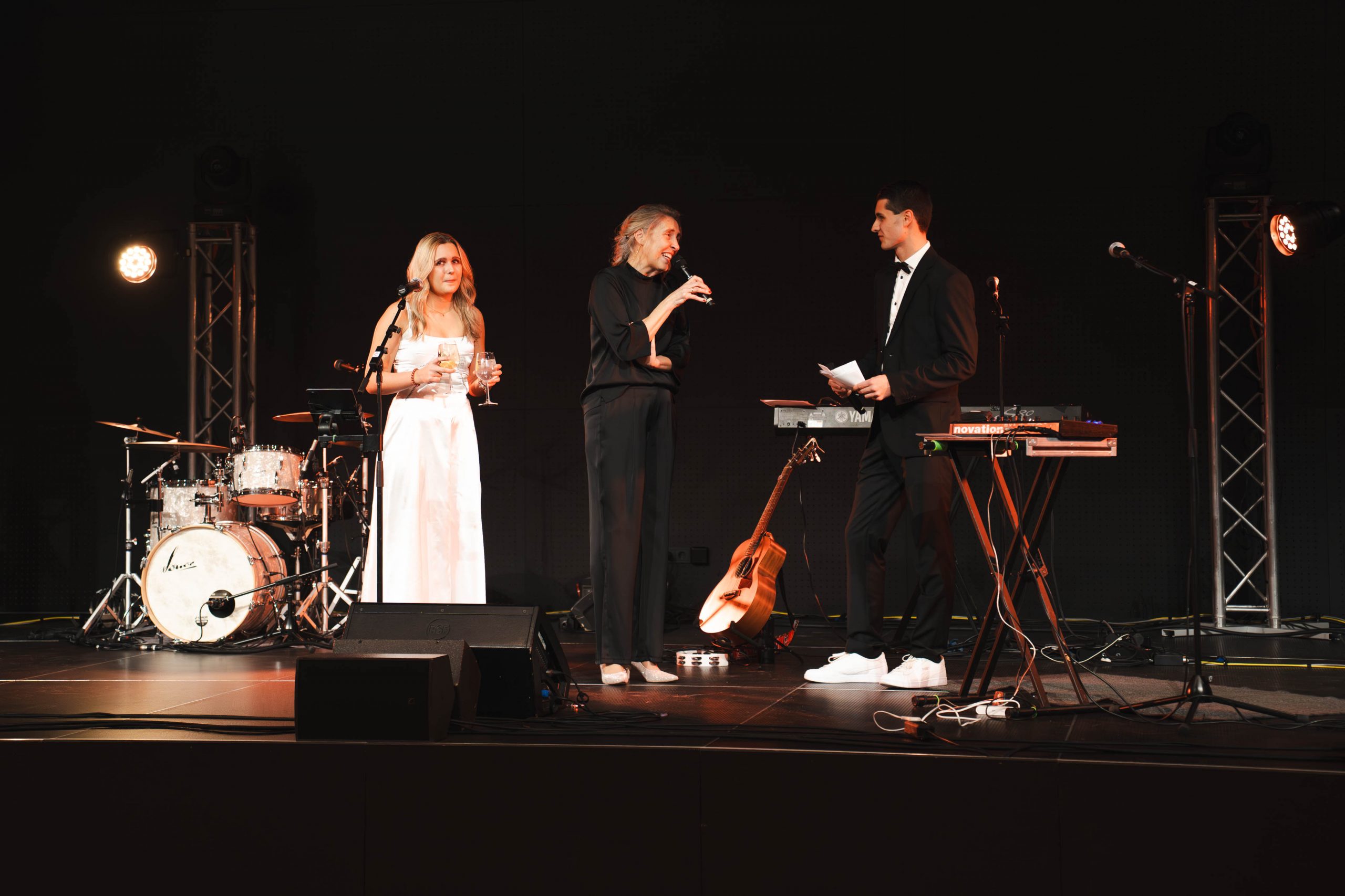 Drei Personen stehen auf einer Bühne mit Musikinstrumenten hinter ihnen. Eine Frau in einem weißen Kleid hält ein Mikrofon, eine andere Person in Schwarz spricht in ein Mikrofon, und ein Mann im Anzug steht in der Nähe und hält ebenfalls ein Mikrofon.