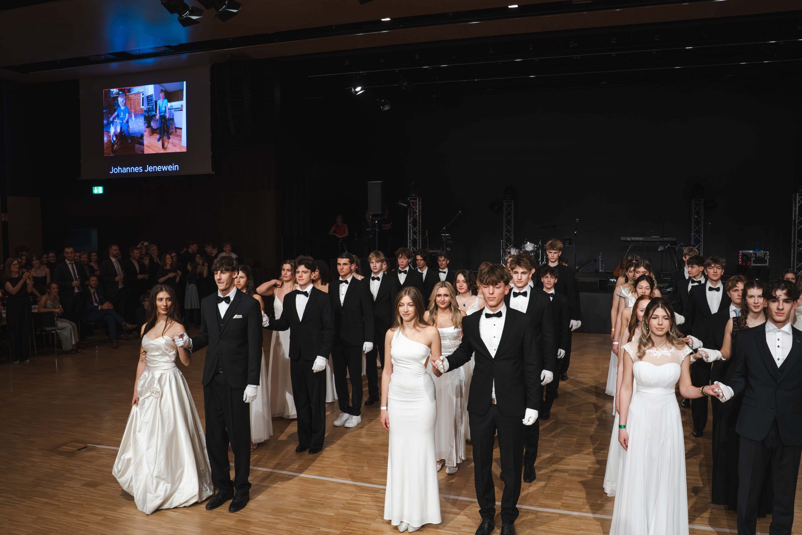 Eine Gruppe junger Männer in schwarzen Smokings und Frauen in weißen Kleidern stehen paarweise auf dem Boden eines Ballsaals und bereiten sich auf einen offiziellen Tanz vor. Ein Publikum schaut zu, und an der Rückwand ist ein projiziertes Bild zu sehen.