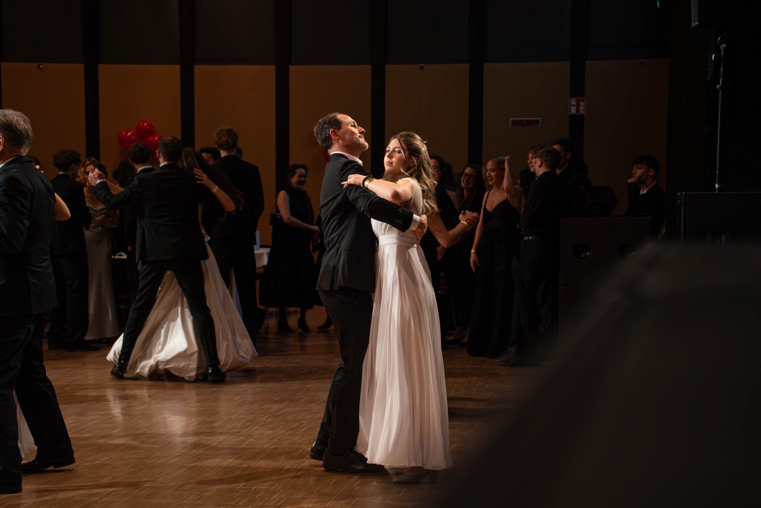 Ein Paar in formeller Kleidung tanzt anmutig auf einem polierten Holzboden in einem Ballsaal, umgeben von anderen Paaren und Schaulustigen in eleganten schwarzen und weißen Kleidern.