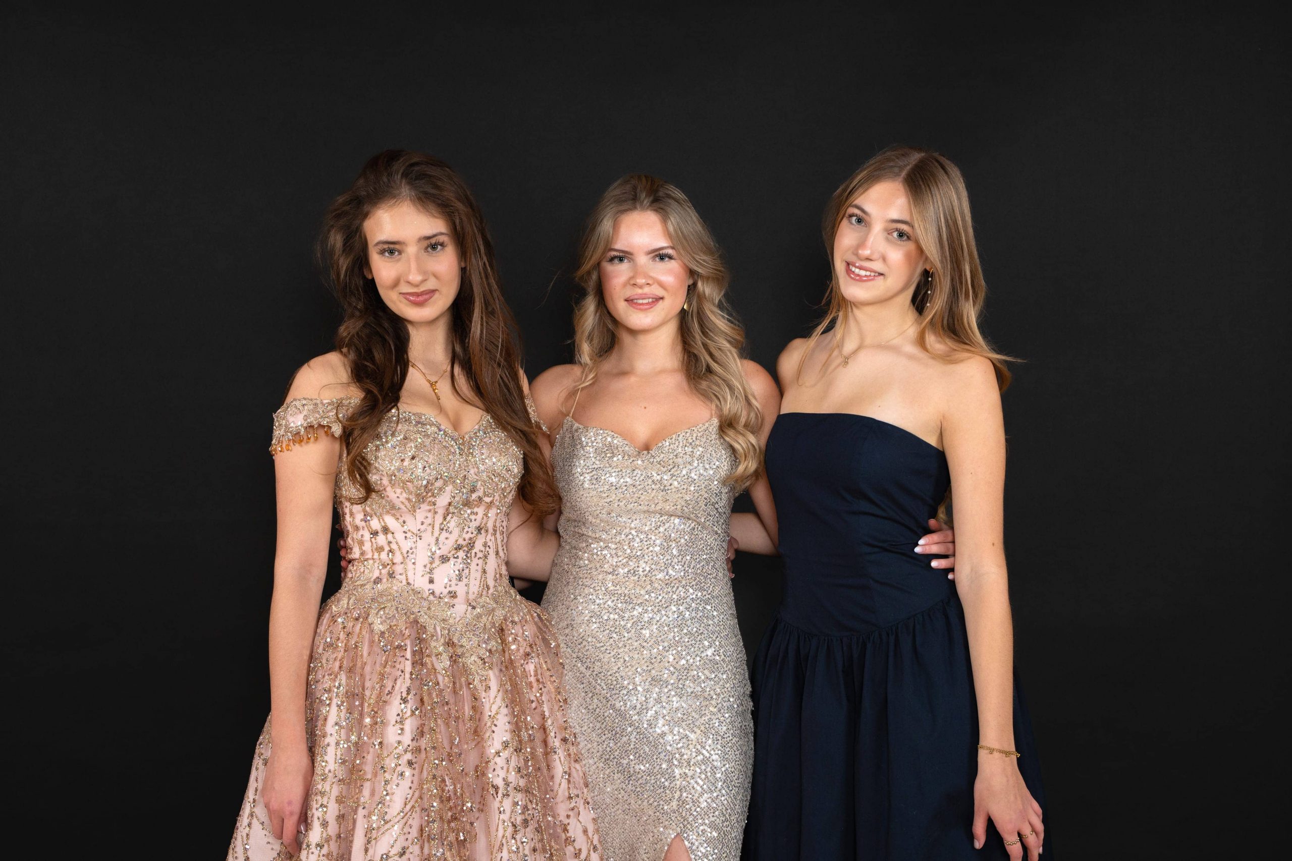 Vor einem schwarzen Hintergrund stehen drei junge Frauen in formellen Abendkleidern zusammen. Eine trägt ein rosa Kleid mit goldenen Details, die zweite ein glitzerndes silbernes Kleid und die dritte ein trägerloses marineblaues Kleid. Sie lächeln.