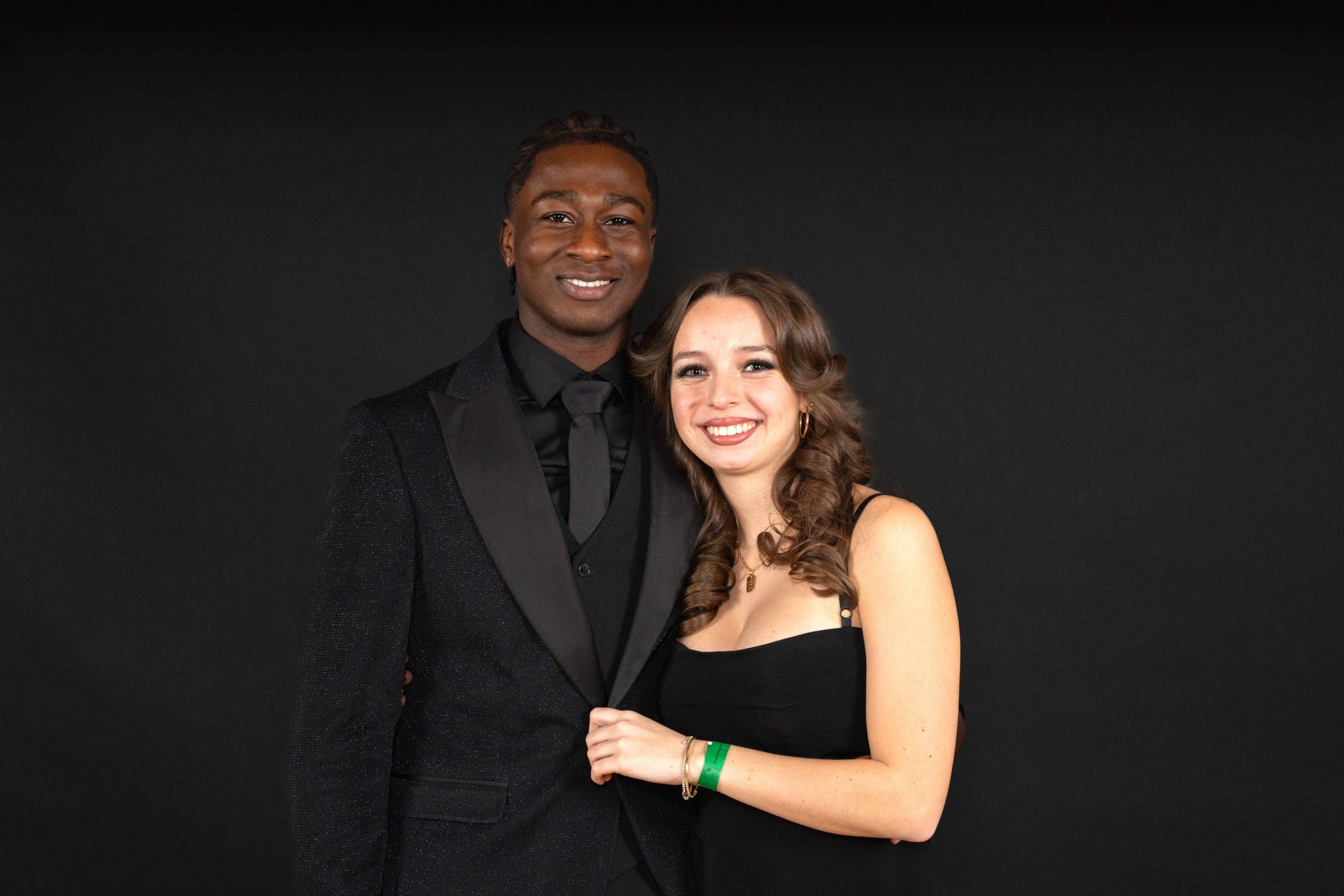 Ein lächelnder Mann in einem schwarzen Anzug und eine Frau in einem schwarzen Kleid stehen zusammen vor einem schlichten schwarzen Hintergrund, posieren eng beieinander und schauen in die Kamera.