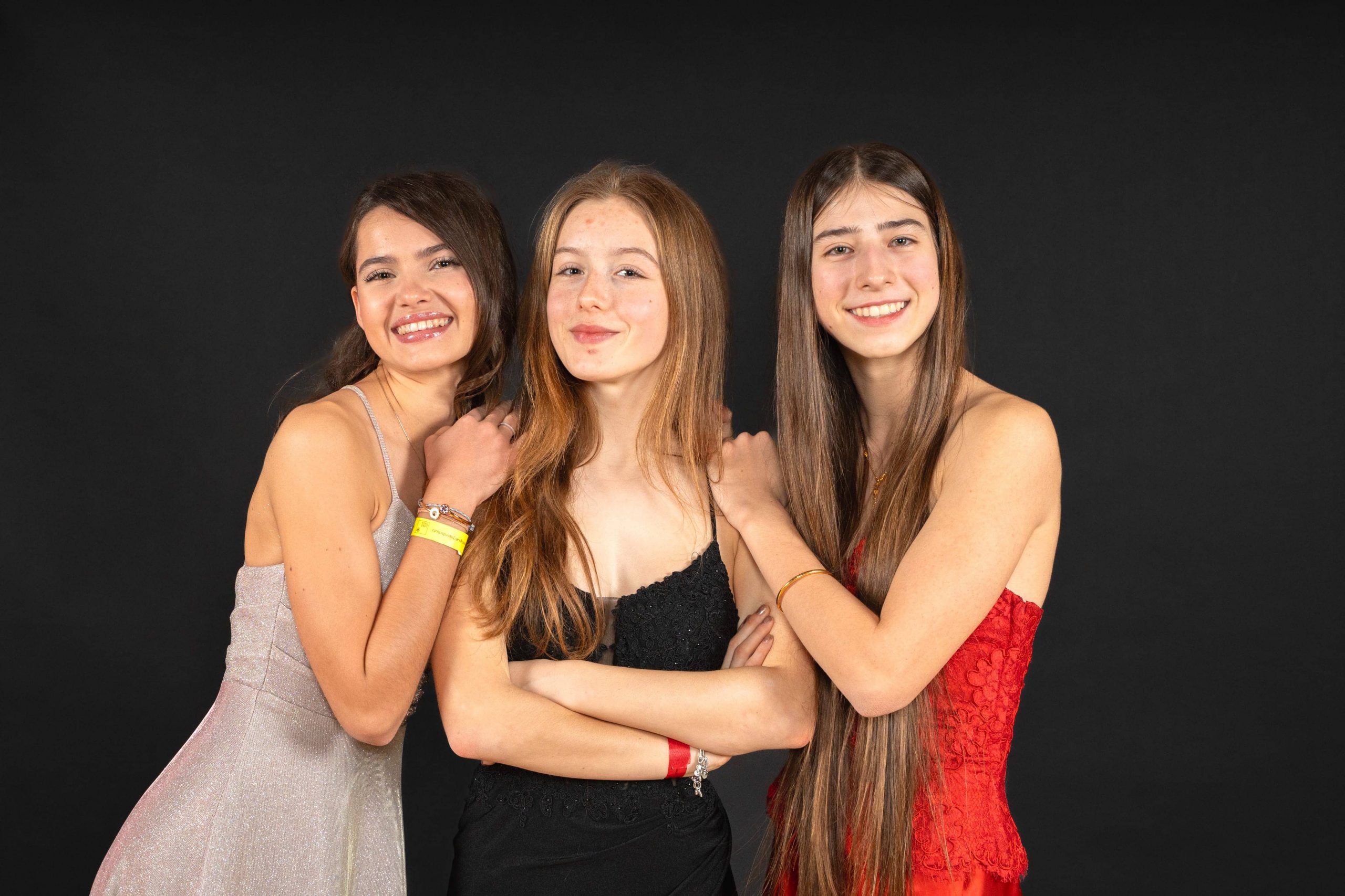 Drei junge Frauen in formellen Kleidern stehen eng beieinander und lächeln vor einem schlichten schwarzen Hintergrund. Eine trägt Silber, eine Schwarz und eine Rot, jede mit langen Haaren, die eine freudige und selbstbewusste Pose einnehmen.