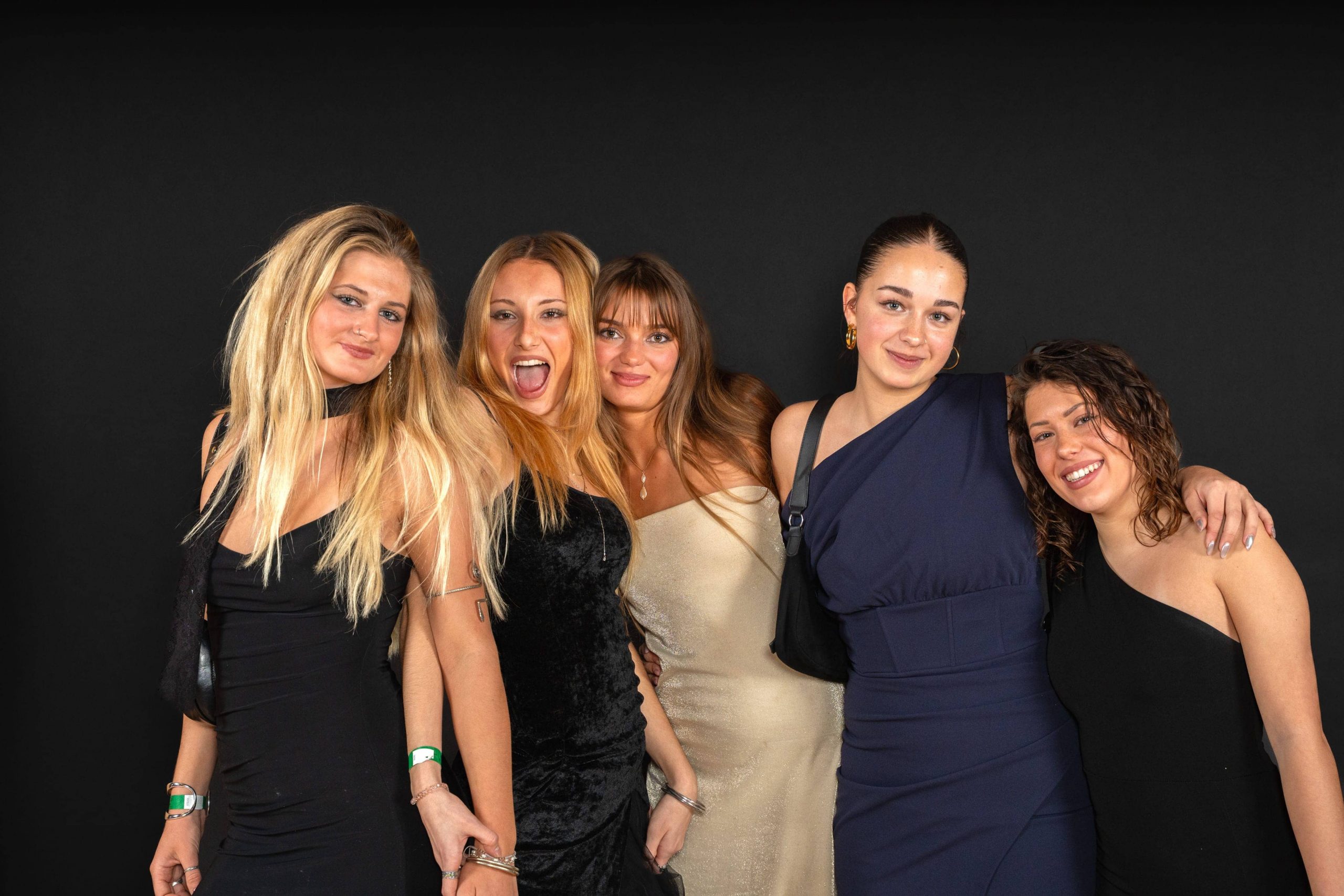 Fünf junge Frauen in eleganten Kleidern stehen dicht beieinander, lächeln und posieren vor einem schlichten schwarzen Hintergrund. Sie haben ihre Arme umeinander gelegt und wirken glücklich und festlich.