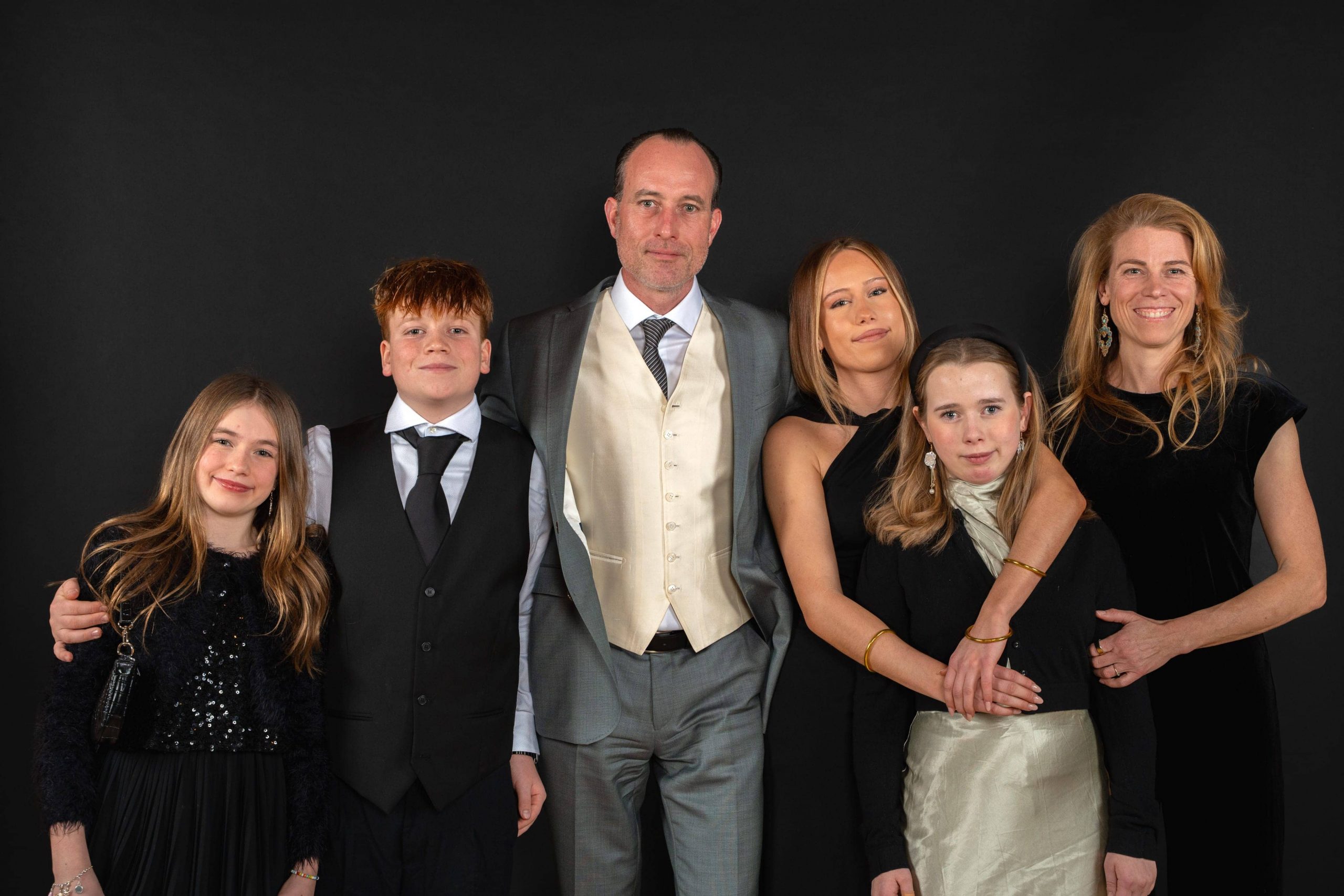 Sechs formell gekleidete Personen stehen dicht beieinander vor einem schwarzen Hintergrund und lächeln in die Kamera. Zu der Gruppe gehören drei Frauen, zwei Mädchen und ein Mann in Anzug und Krawatte.