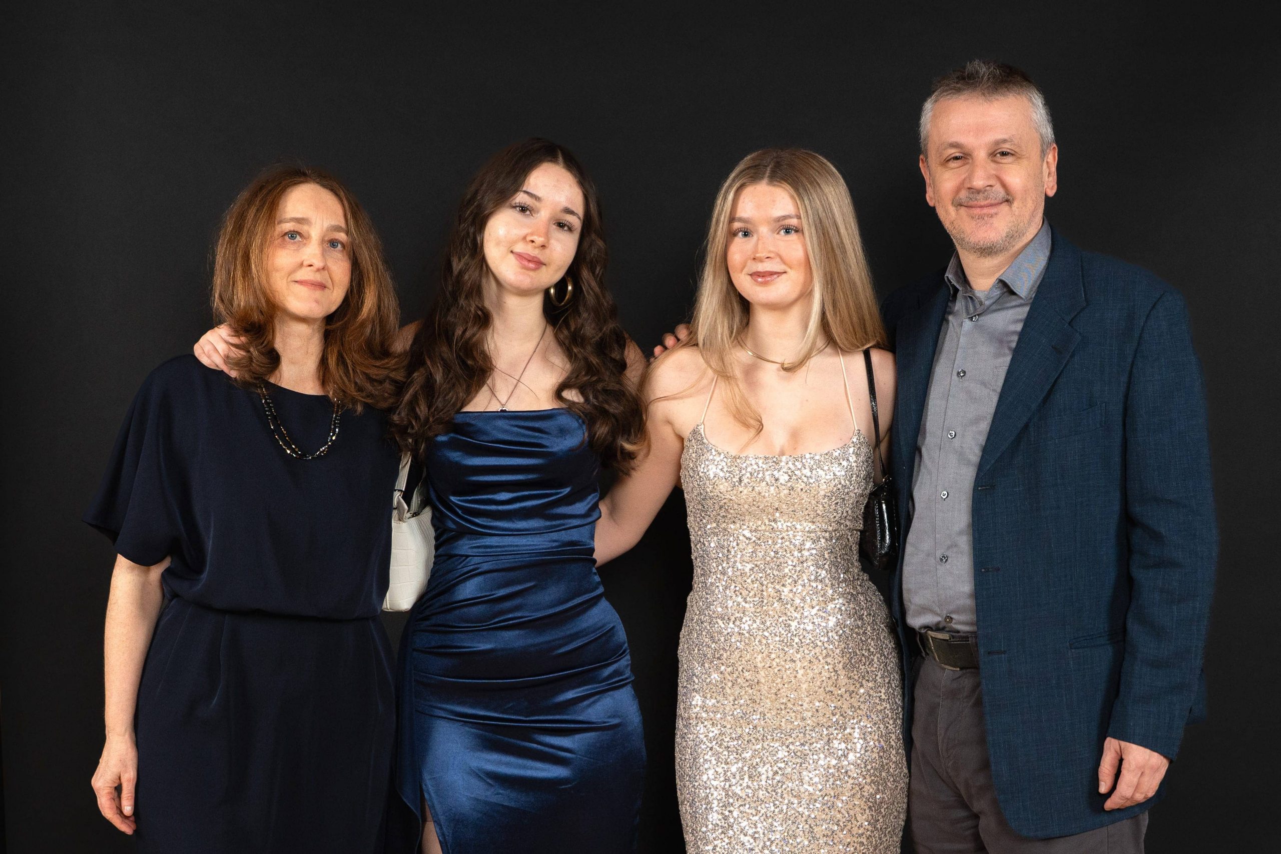 Vier Personen stehen in einer Reihe vor einem schwarzen Hintergrund und lächeln. Auf der linken Seite stehen zwei Frauen, eine in einem marineblauen Kleid, die andere in einem glänzenden blauen Kleid. Auf der rechten Seite stehen eine Frau in einem glitzernden goldenen Kleid und ein Mann in einem blauen Anzug.
