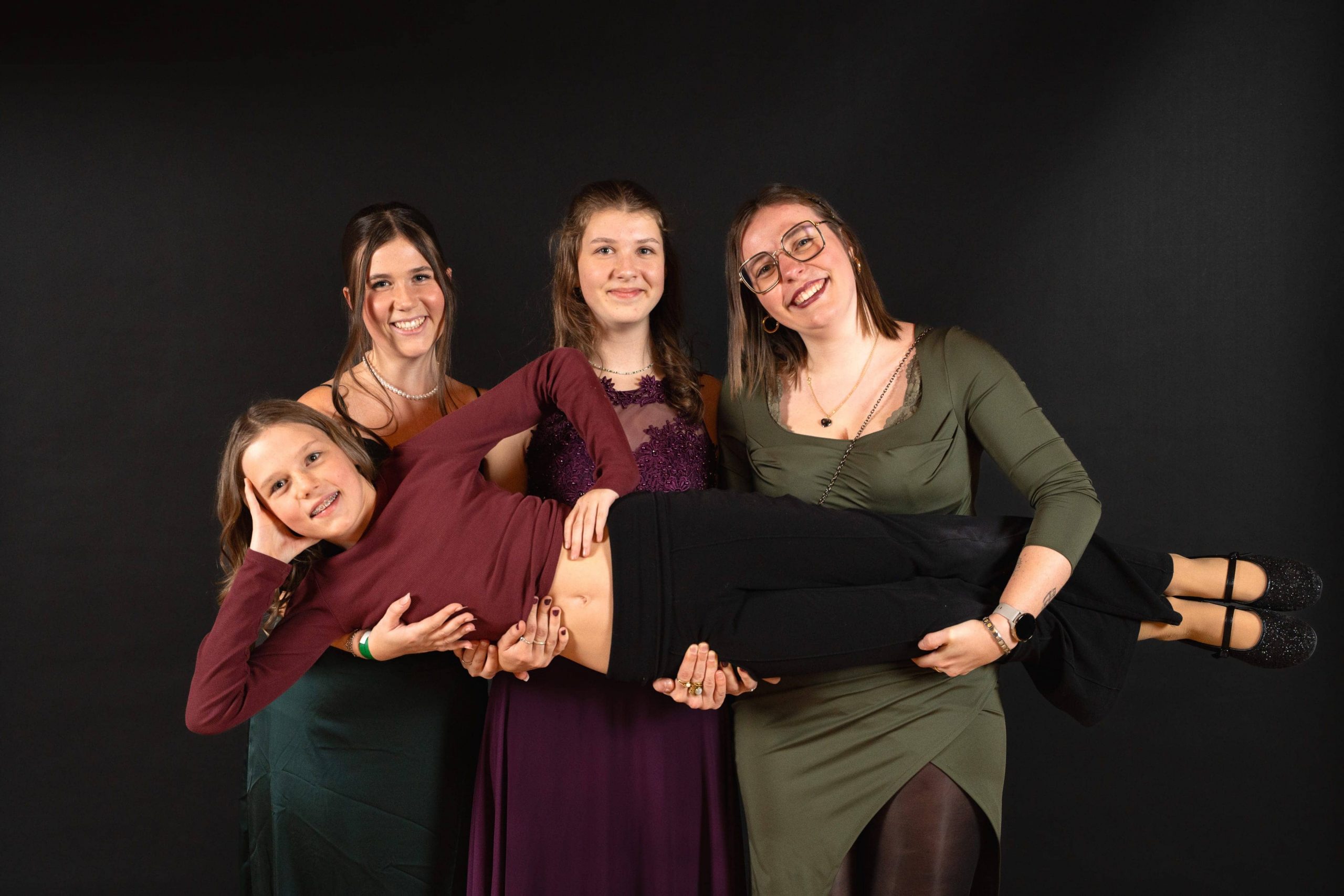 Vier lächelnde junge Frauen posieren gemeinsam vor einem schwarzen Hintergrund. Drei von ihnen stehen und halten die vierte hoch, die waagerecht vor ihnen liegt, ein kastanienbraunes Top trägt und lächelnd die Hand an den Kopf hält.