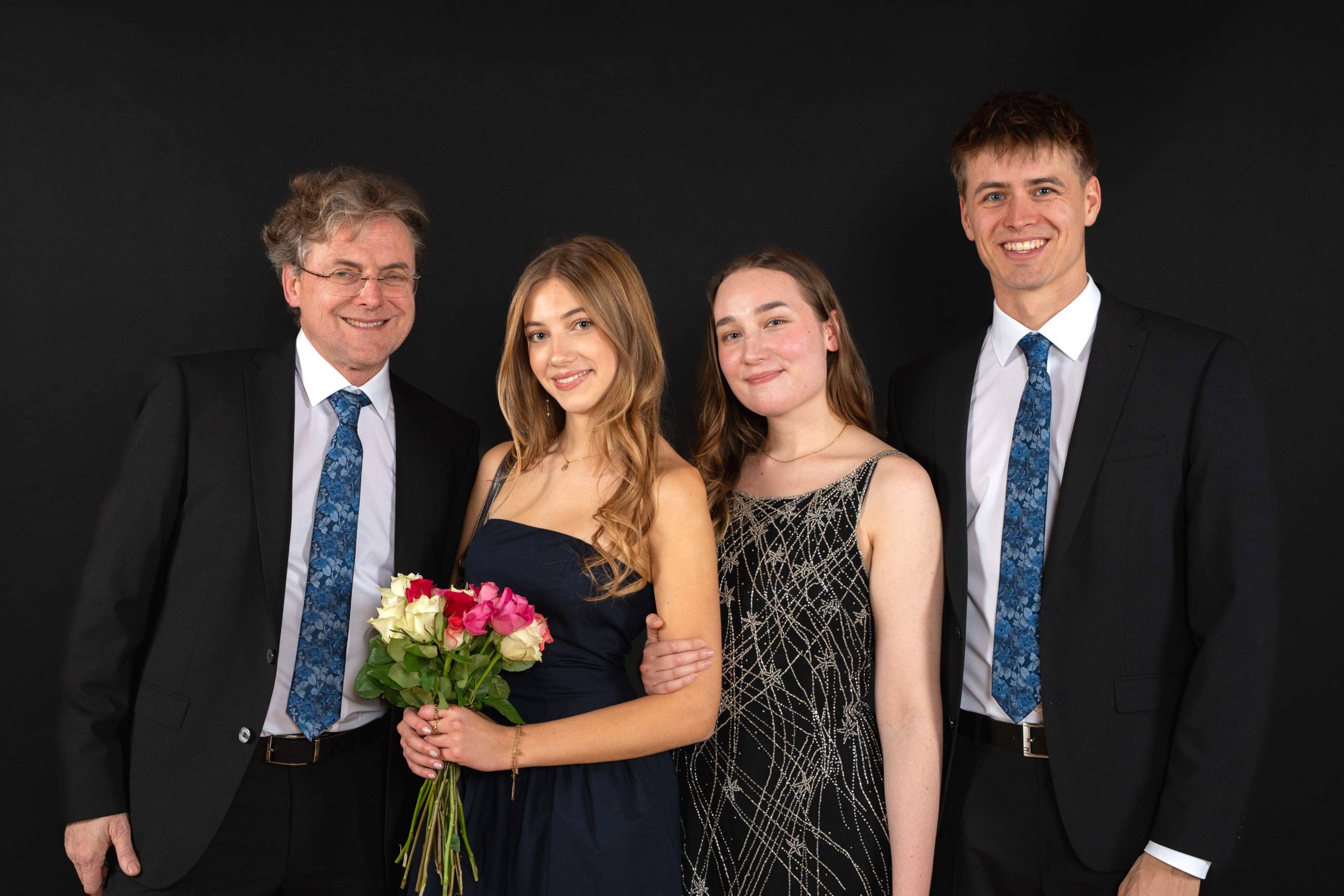 Vier förmlich gekleidete Personen posieren vor einem schwarzen Hintergrund. Zwei Männer in Anzügen stehen auf beiden Seiten von zwei Frauen; eine Frau hält einen Strauß mit rosa und weißen Blumen. Alle lächeln in die Kamera.