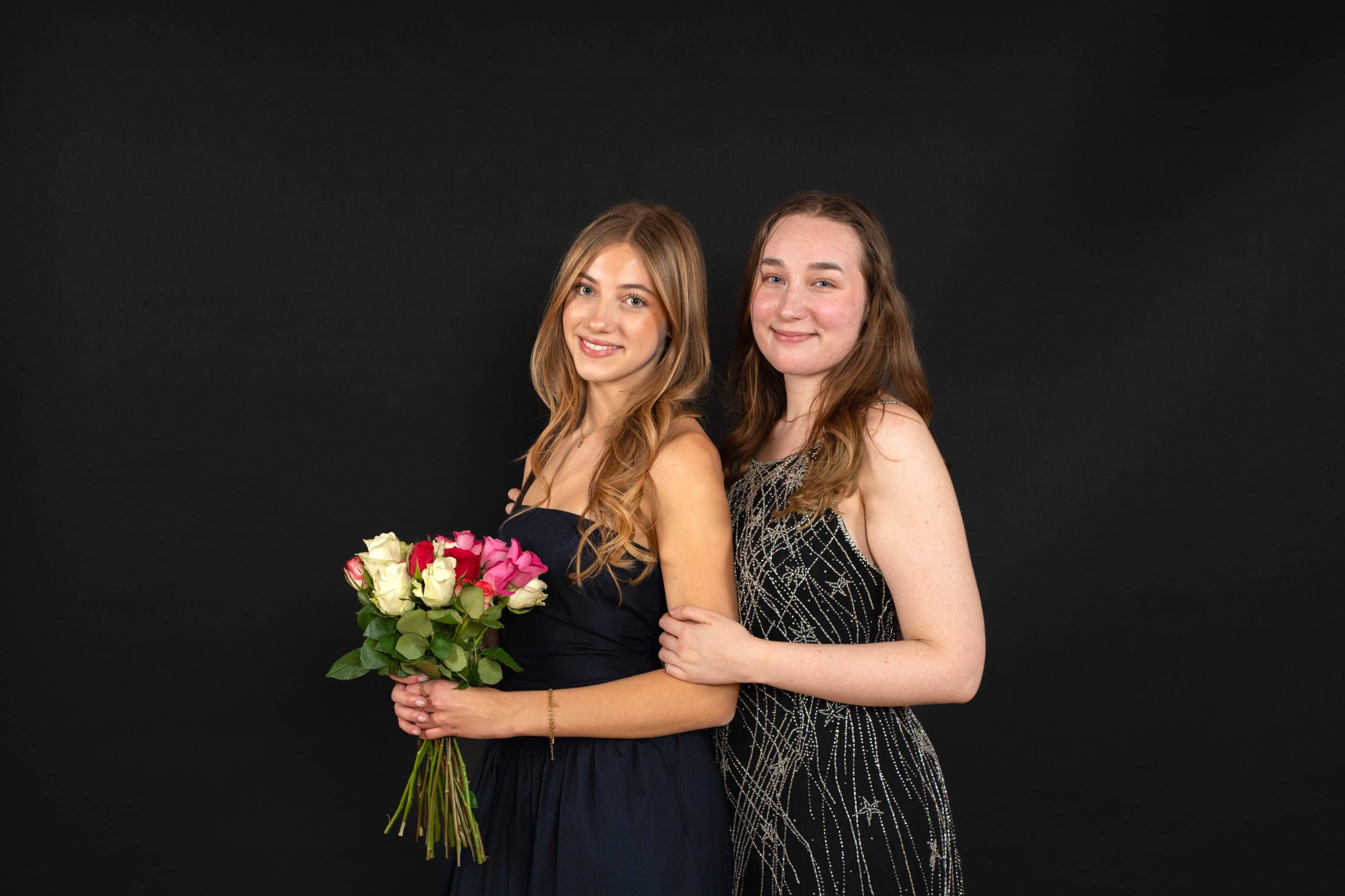 Zwei junge Frauen stehen dicht nebeneinander vor einem schwarzen Hintergrund. Eine hält einen Strauß roter und weißer Rosen in der Hand, beide lächeln und tragen elegante formelle Kleider.