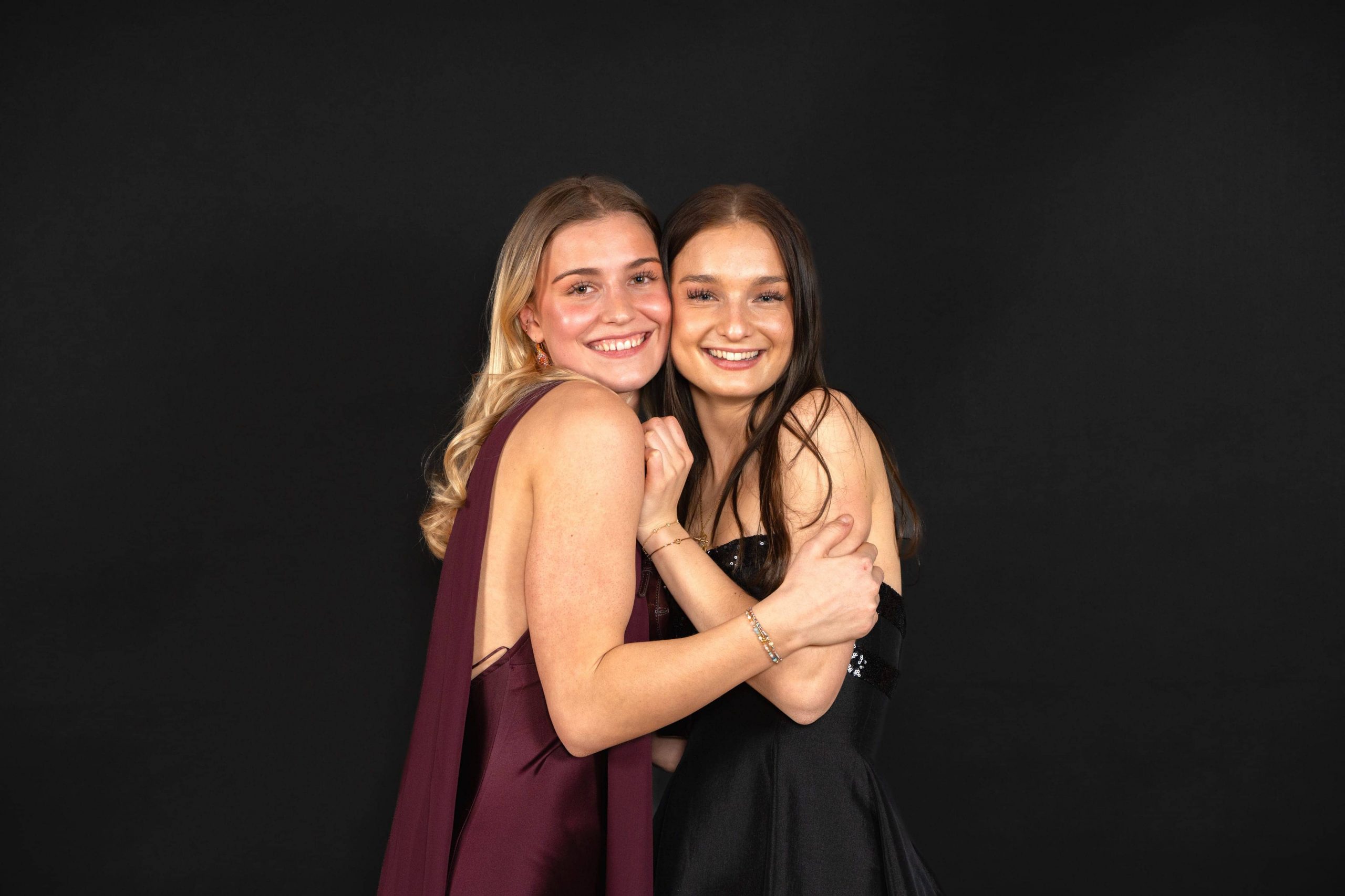 Zwei junge Frauen in formellen Kleidern stehen eng beieinander, lächeln und umarmen sich vor einem schlichten schwarzen Hintergrund.