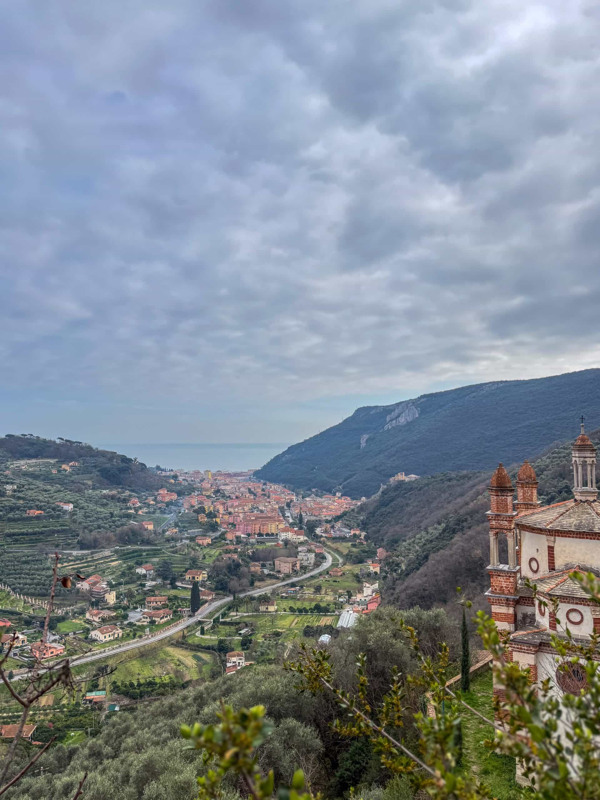 Blick auf eine kleine Stadt in einem grünen Tal, mit gewundenen Straßen, verstreuten Häusern und einer großen Kirche im Vordergrund. Zu beiden Seiten erheben sich Hügel und Berge unter einem wolkenverhangenen Himmel, und in der Ferne ist das Meer zu sehen.