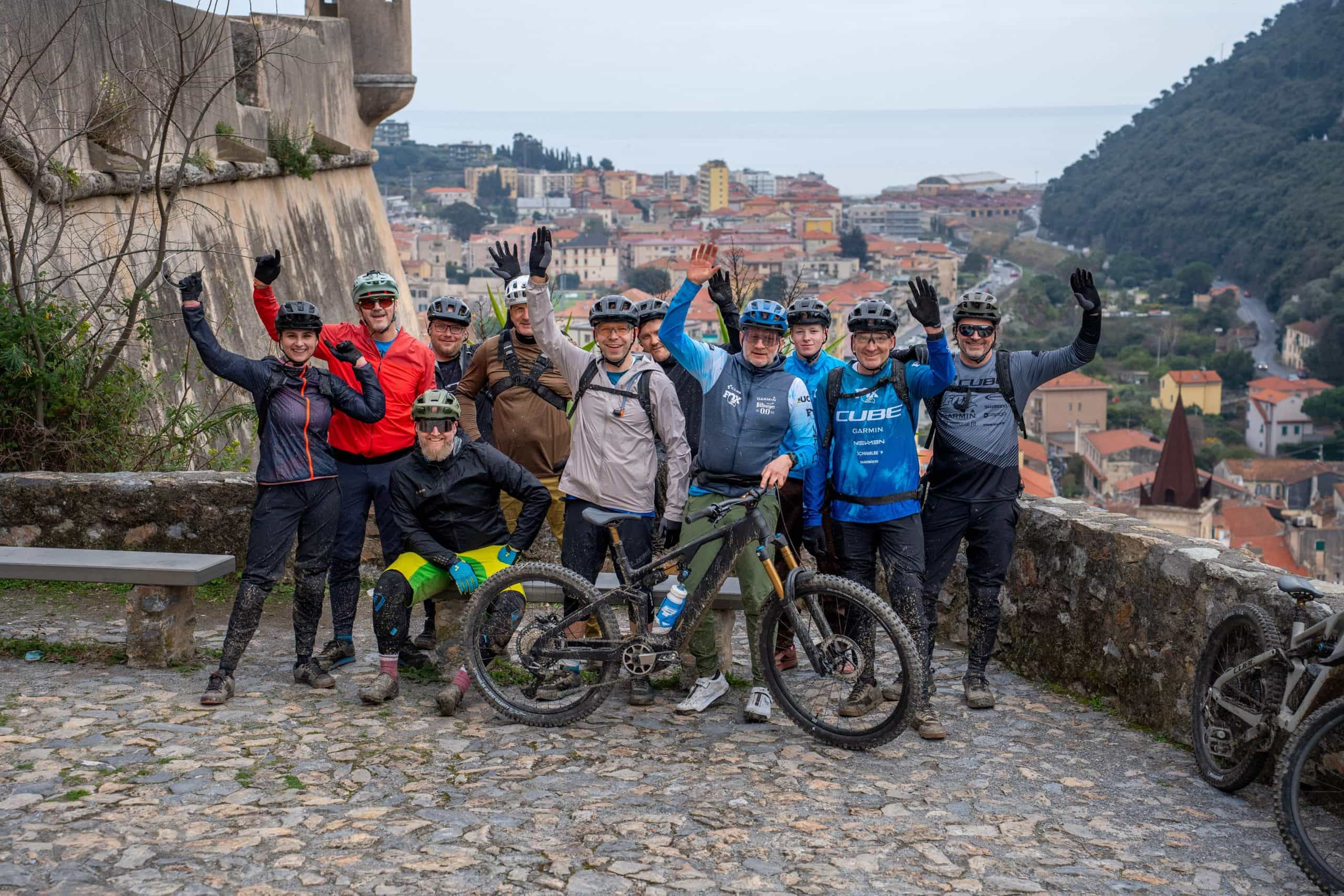 Eine Gruppe von Radfahrern mit Helmen und Ausrüstung posiert lächelnd und winkend auf einem Steinweg mit Blick auf eine Stadt mit Hügeln und dem Meer im Hintergrund. Vor ihnen steht ein Mountainbike.
