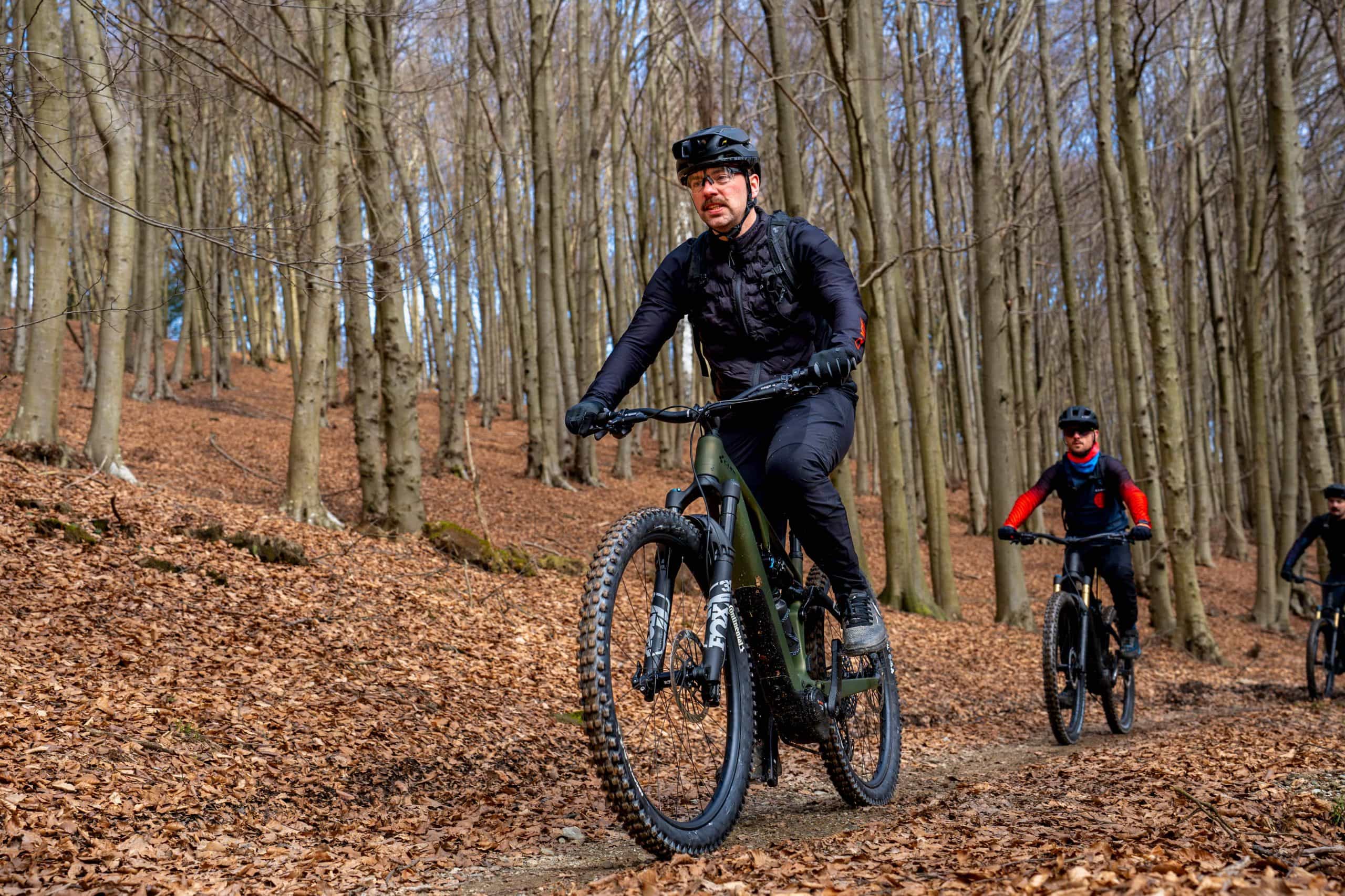 Drei Radfahrer fahren mit Mountainbikes auf einem mit trockenem Laub bedeckten Weg in einem blattlosen Wald, sie tragen Helme und dunkle Fahrradkleidung. Die Bäume im Hintergrund lassen vermuten, dass es Herbst oder Vorfrühling ist.