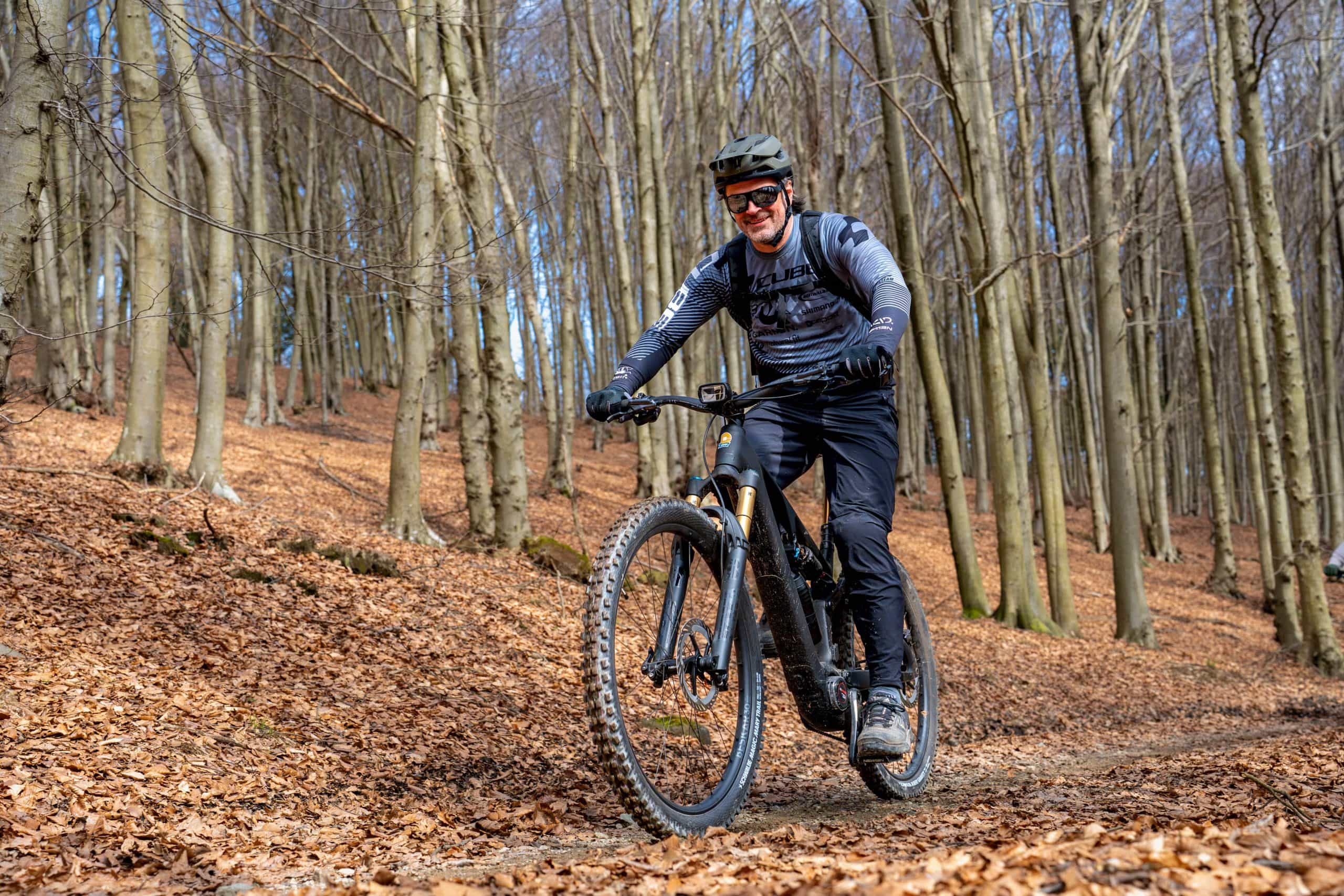 Ein Mann, der einen Helm, eine Sonnenbrille und eine Outdoor-Ausrüstung trägt, fährt an einem sonnigen Tag mit seinem Mountainbike auf einem mit Laub bedeckten Weg durch einen Wald mit hohen, kahlen Bäumen.