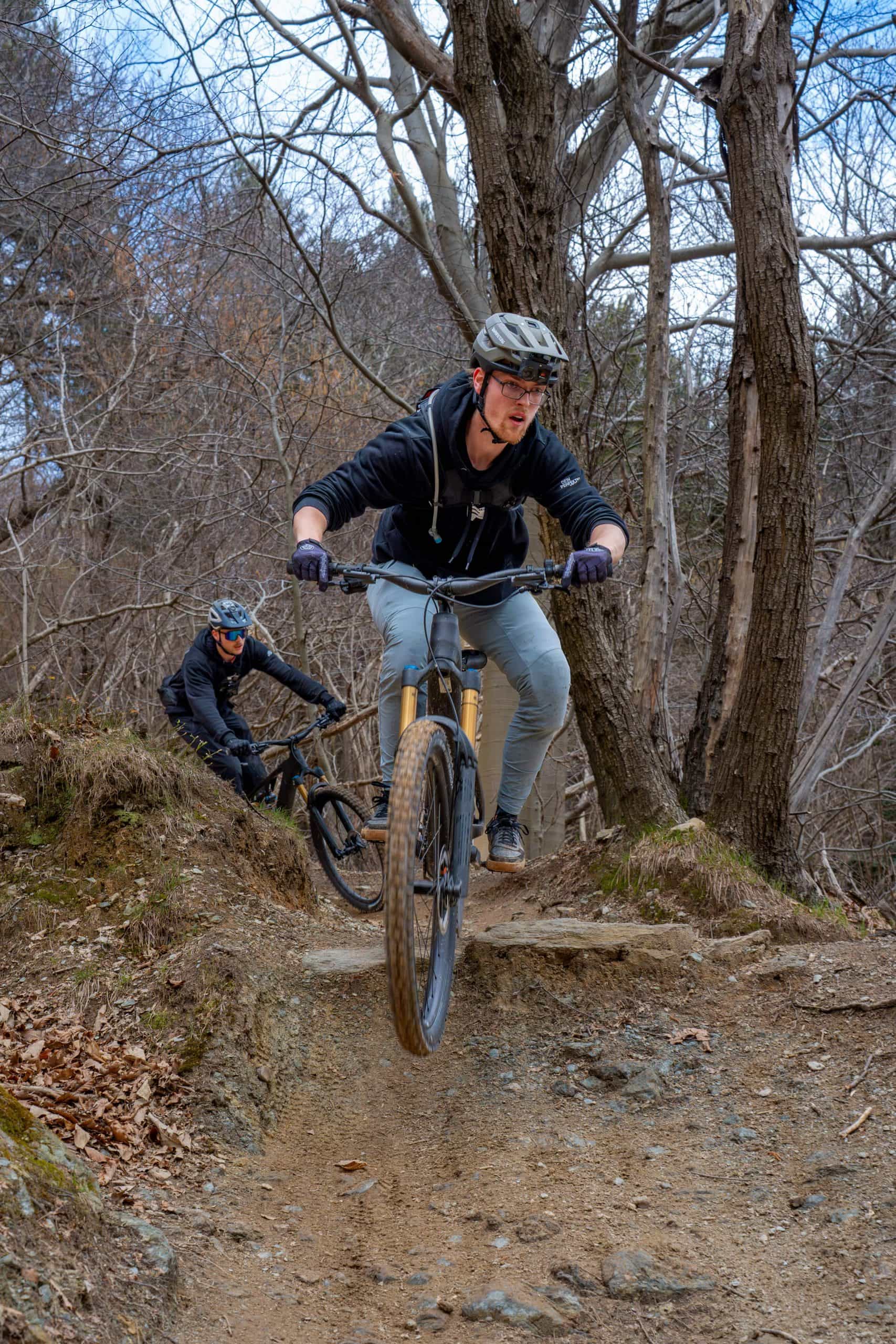 Zwei Personen mit Helmen fahren mit Mountainbikes auf einem unbefestigten Weg in einem Wald. Der vordere Fahrer springt von einem kleinen Abhang in die Luft, während der zweite Fahrer dicht hinter ihm folgt. Kahle Bäume umgeben den Weg.