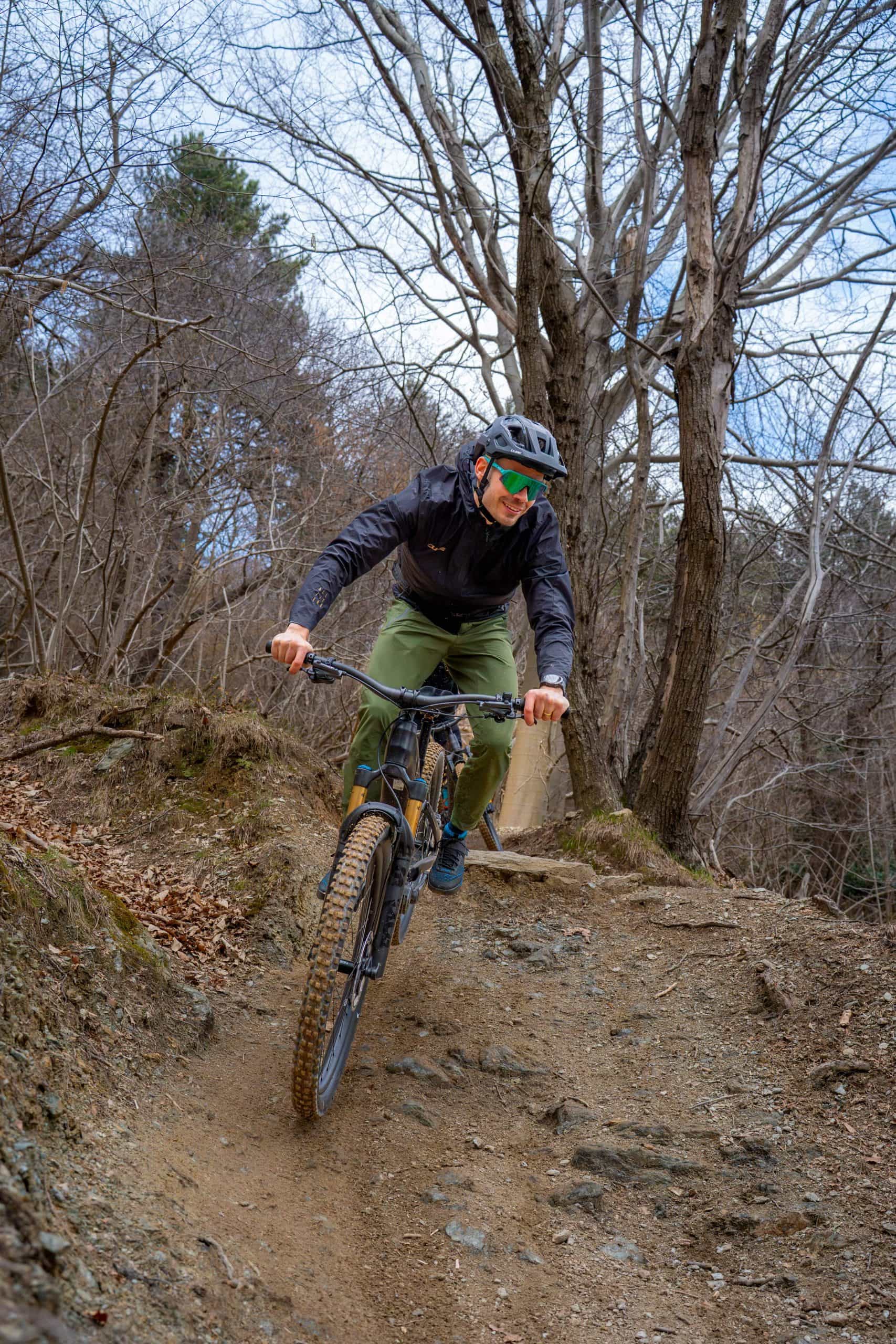 Eine Person, die einen Helm, eine Sonnenbrille und eine Outdoor-Ausrüstung trägt, fährt mit einem Mountainbike auf einem holprigen, unbefestigten Weg durch einen Wald mit kahlen Bäumen.