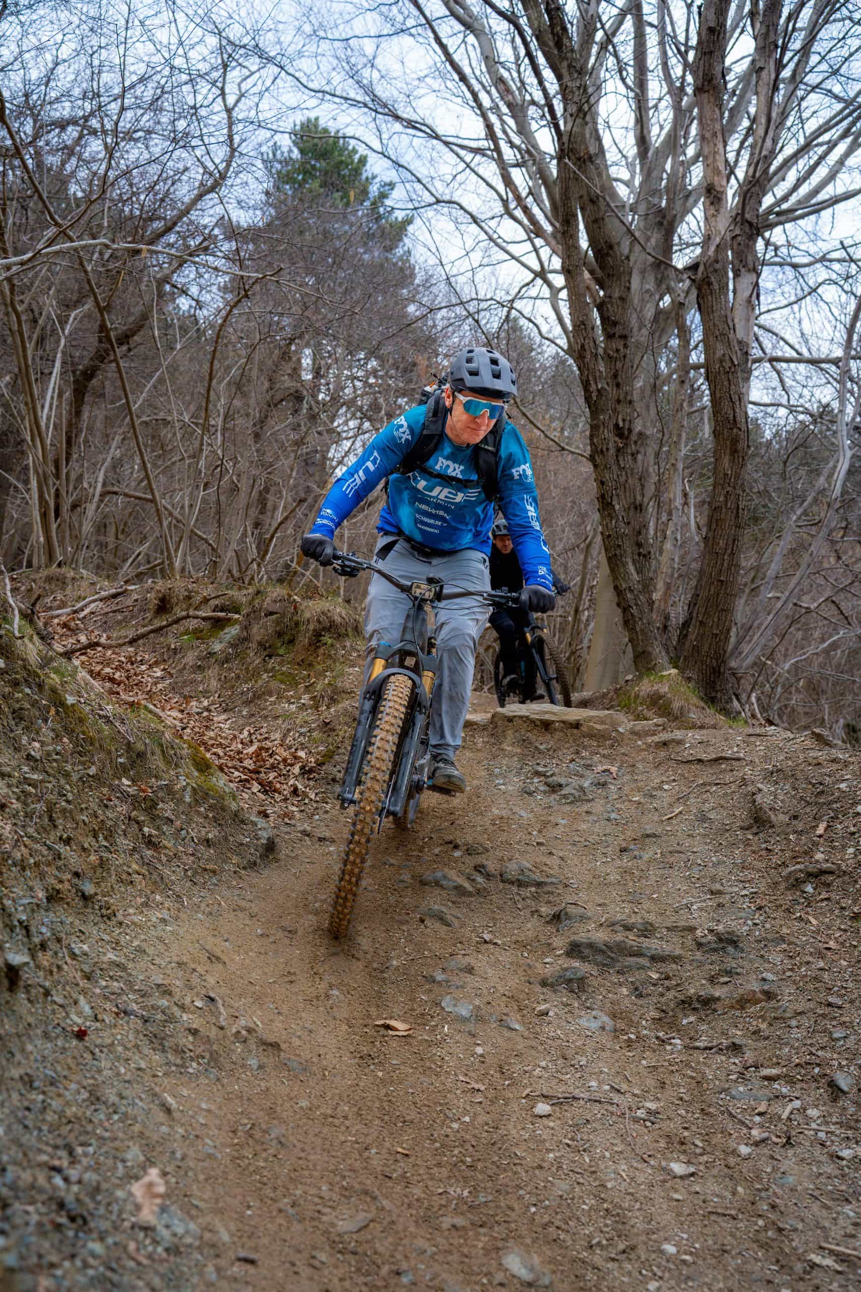 Eine Person, die eine blaue Jacke, einen Helm und eine Sonnenbrille trägt, fährt mit einem Mountainbike auf einem unbefestigten Weg durch einen Wald mit kahlen Bäumen. Ein anderer Radfahrer folgt hinter ihr.