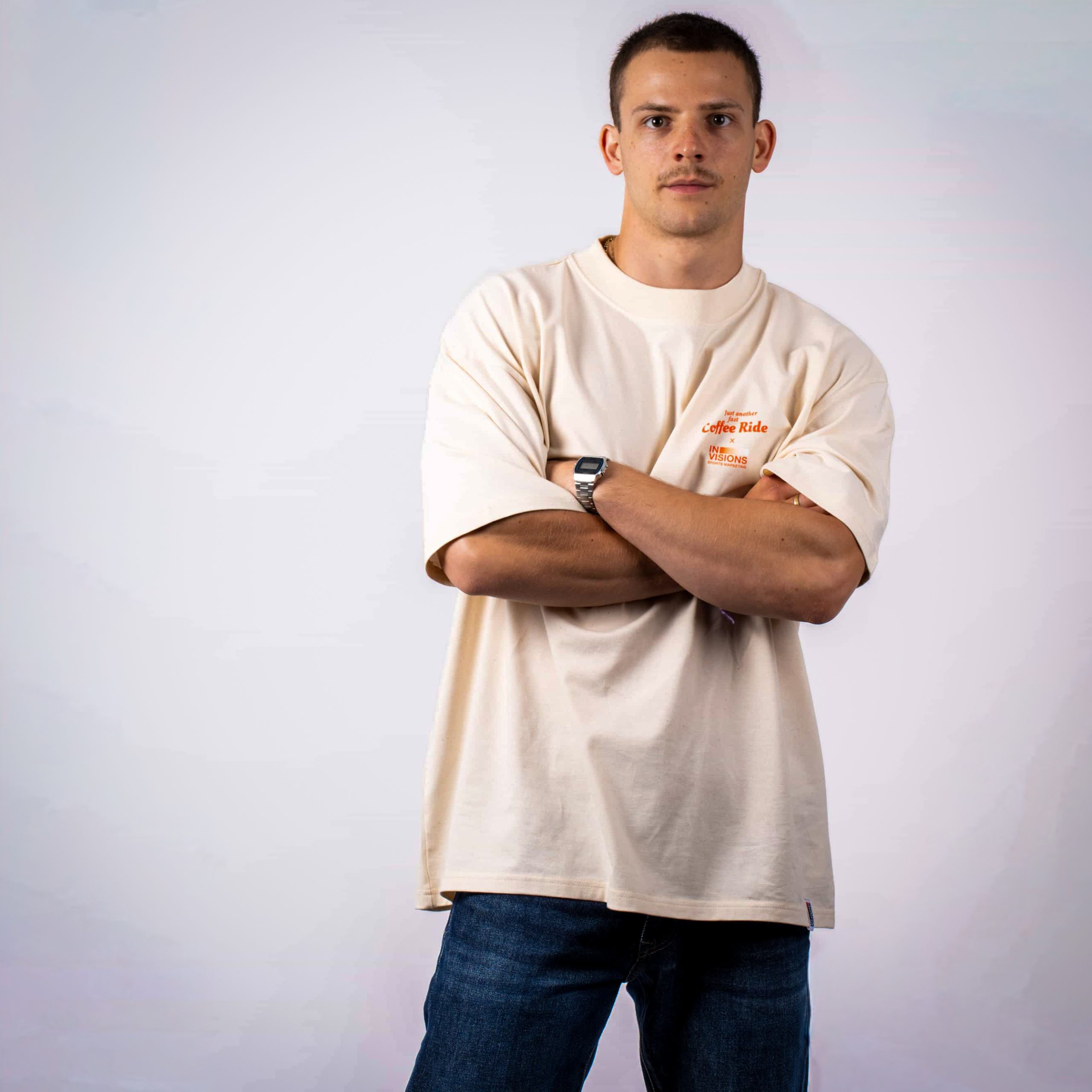 Ein Mann steht mit verschränkten Armen vor einem schlichten weißen Hintergrund, trägt ein beigefarbenes übergroßes T-Shirt, blaue Jeans und eine Uhr am linken Handgelenk.