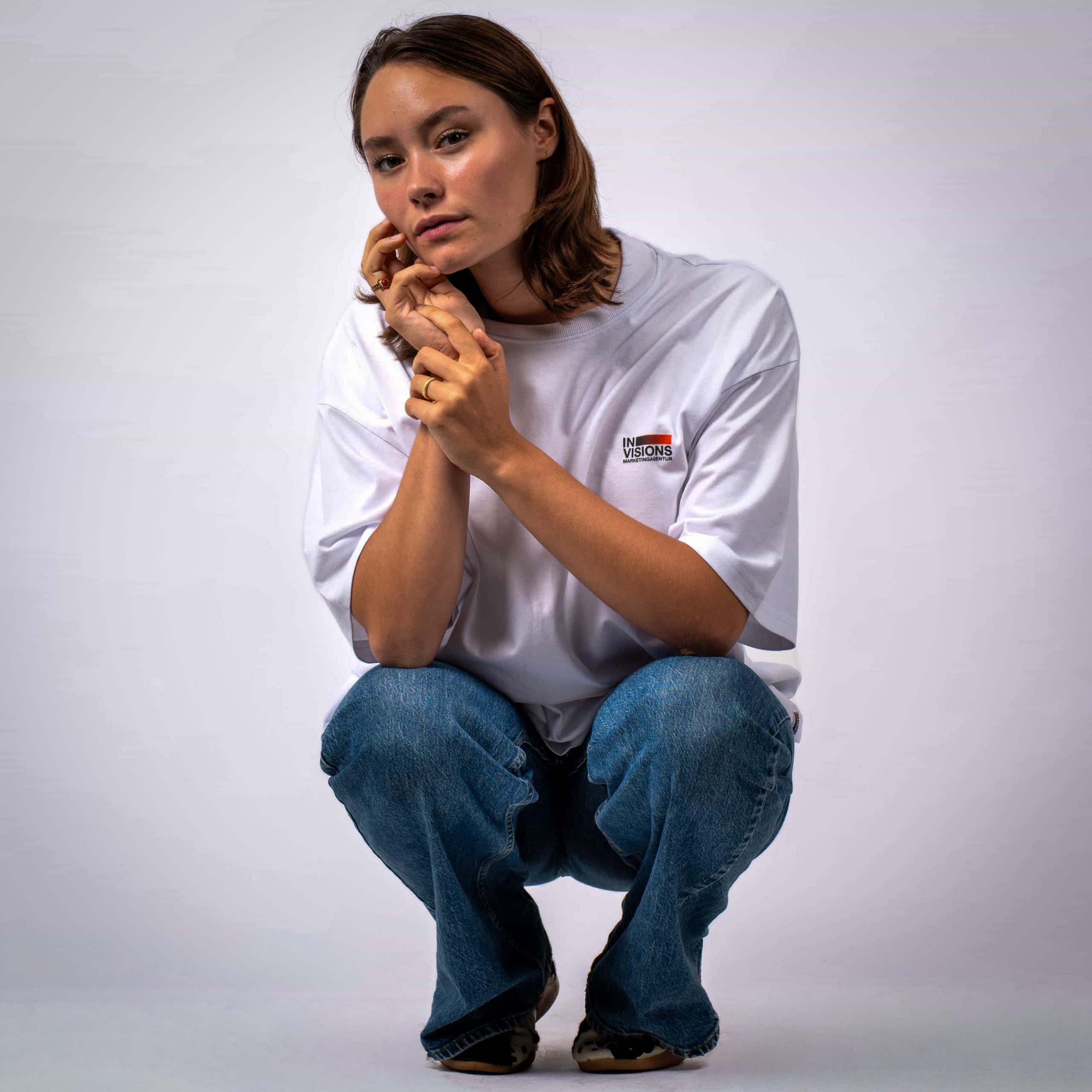 Eine Frau mit braunen Haaren hockt vor einem schlichten weißen Hintergrund und trägt ein weißes T-Shirt und blaue Jeans. Sie blickt mit neutraler Miene nach vorne und stützt ihr Kinn auf ihre Hand.