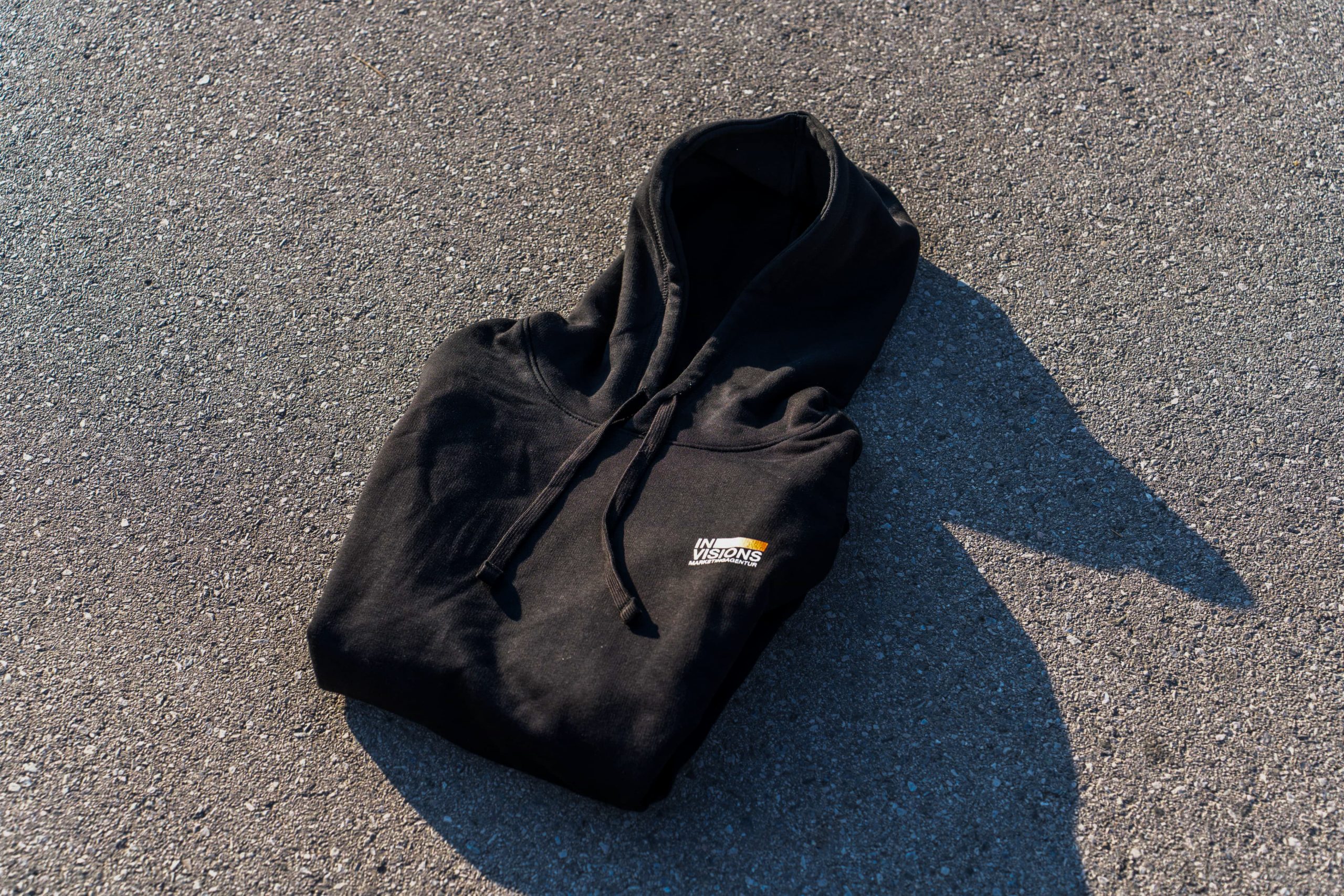 Ein schwarzes Kapuzensweatshirt mit einem kleinen weiß-orangenen Logo auf der Brust ist ordentlich gefaltet und liegt auf einer sonnenbeschienenen Asphaltfläche, die einen Schatten nach rechts wirft.