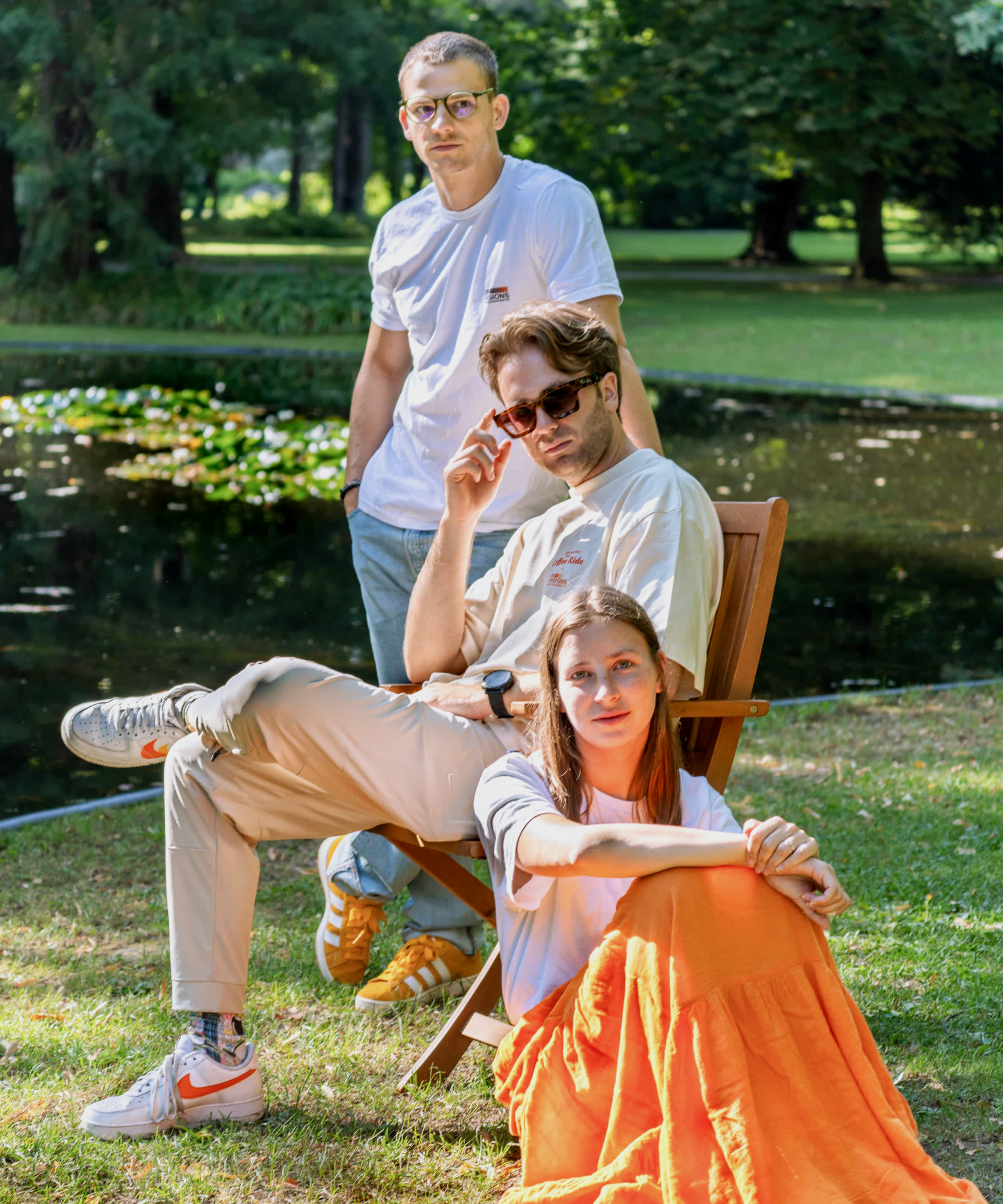 Drei Personen posieren im Freien an einem Teich. Eine sitzt auf einem Stuhl und trägt eine Sonnenbrille, eine andere sitzt in einem orangefarbenen Rock im Gras, und die dritte steht hinter ihnen. Bäume und Grünflächen umgeben sie im Hintergrund.