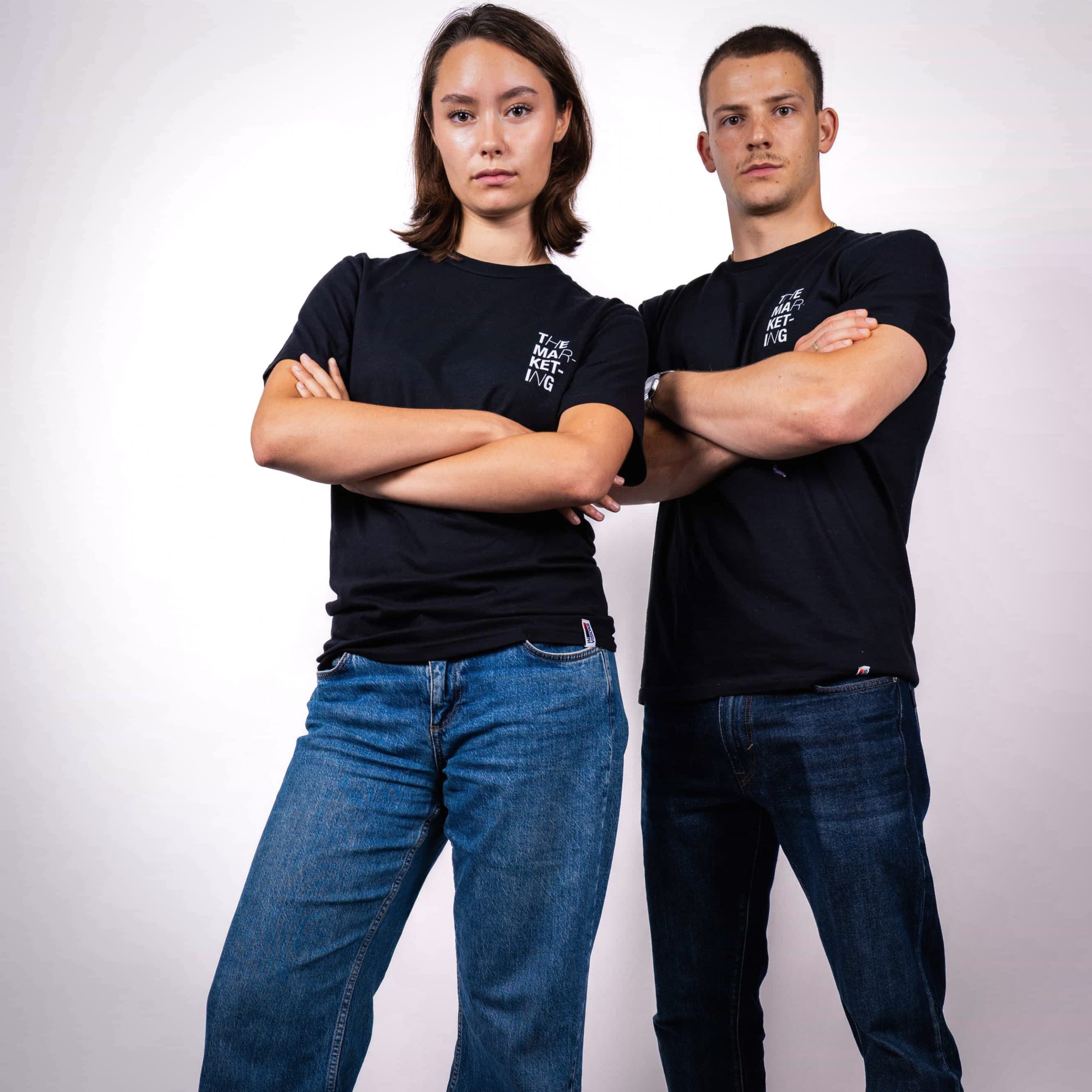 Zwei Personen stehen selbstbewusst mit verschränkten Armen da, tragen schwarze T-Shirts und blaue Jeans. Die Person auf der linken Seite trägt Schuhe mit Kuhaufdruck, während die Person auf der rechten Seite gelbe Turnschuhe trägt. Beide blicken nach vorne vor einem schlichten weißen Hintergrund.
