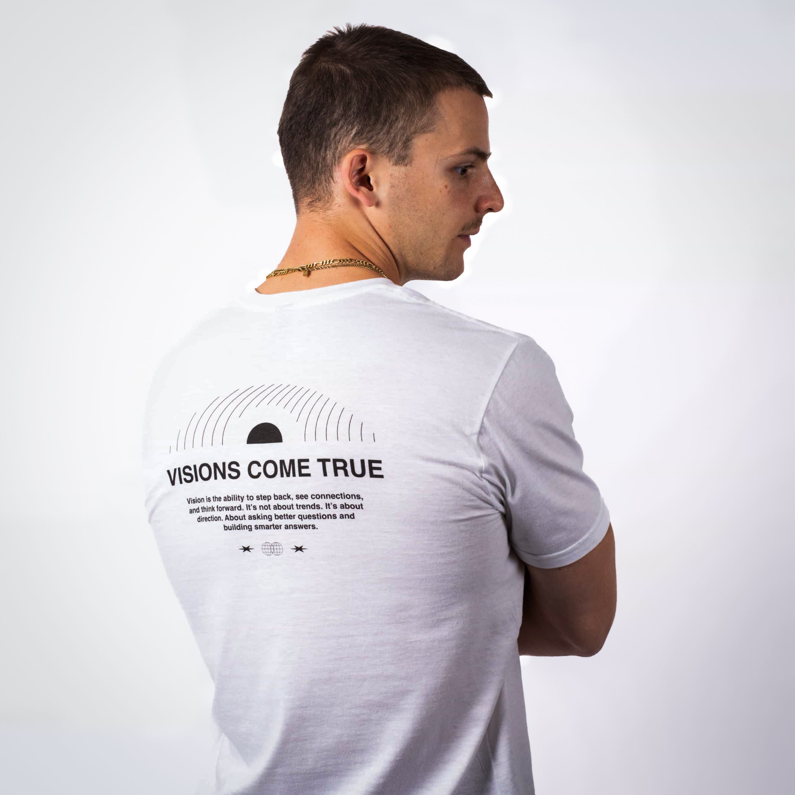 Ein Mann in blauen Jeans und einem weißen T-Shirt steht mit dem Rücken zur Kamera. Auf dem T-Shirt sind der Satz "VISIONS COME TRUE" und ein Text auf der Rückseite aufgedruckt. Der Hintergrund ist einfarbig weiß.