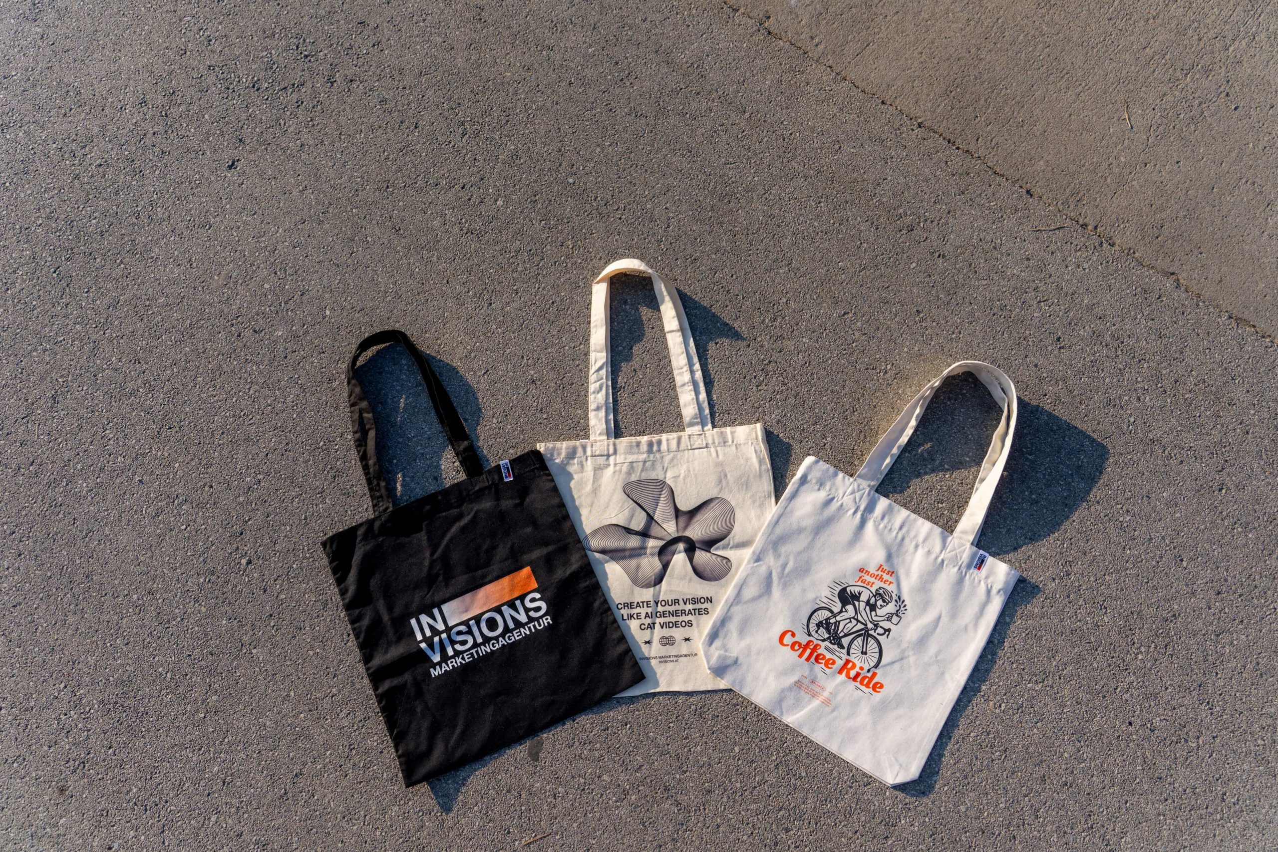Drei Tragetaschen sind auf einer gepflasterten Fläche ausgelegt. Die linke ist schwarz mit dem Text "IN VISIONS", die mittlere ist beige mit einem geometrischen Muster, und die rechte ist beige mit einem Cartoon-Radfahrer und dem Text "Coffee Ride".